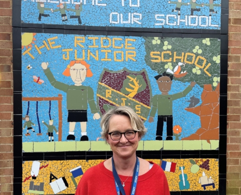 Image shows Beverly Furbur from The Ridge Primary school smiling infront of a sign wearing a red top