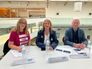 Judging panel  of 3 sat smiling at a table, 2 x women and 1 x man in blue jacket